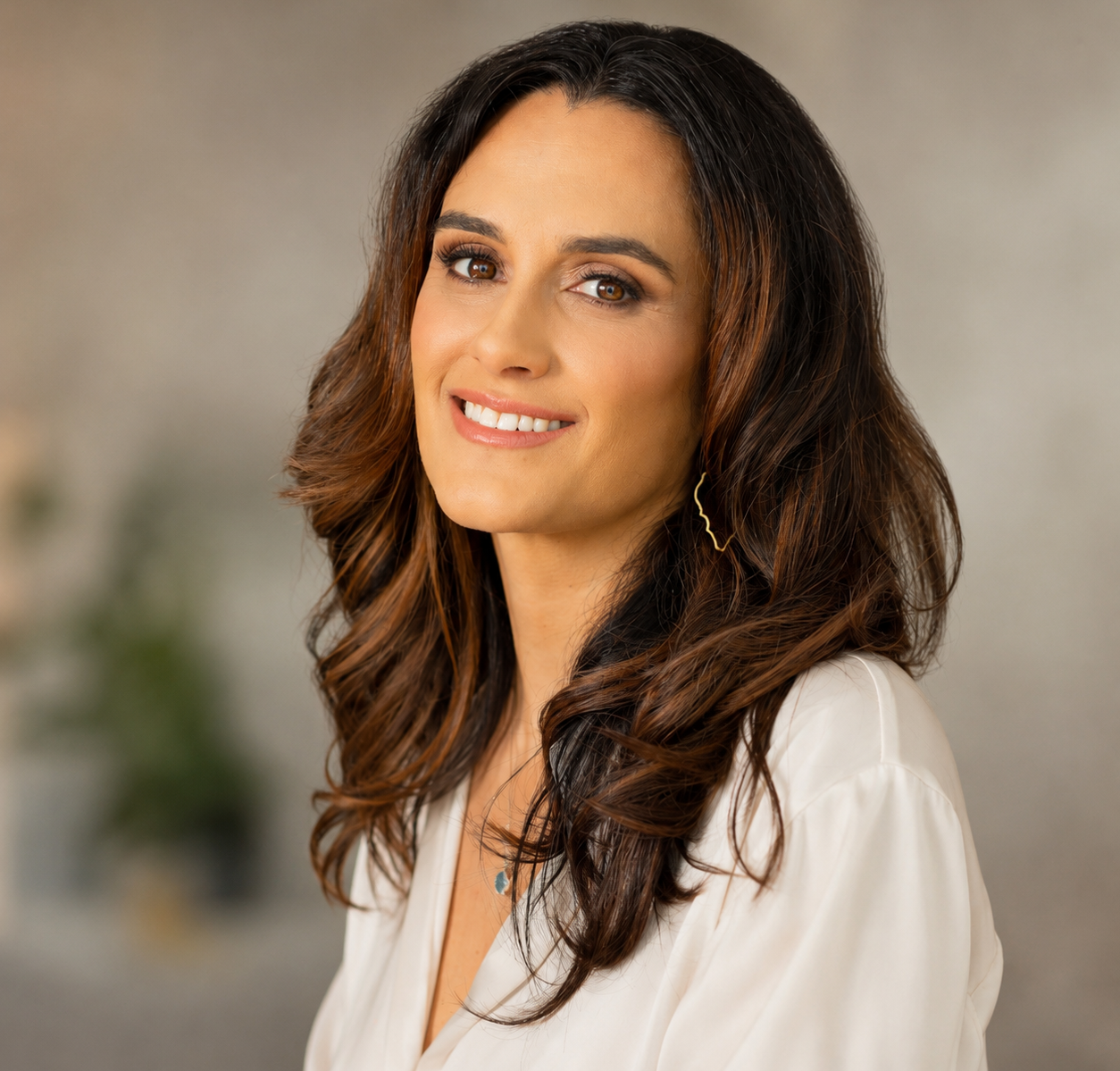 Smiling woman with long dark hair in a white blouse against a blurred indoor background