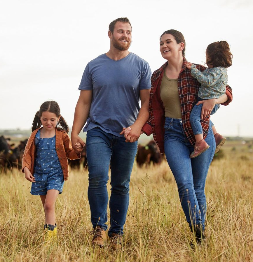 Family walking through a grassy field, smiling and holding hands, with two children and a dog.