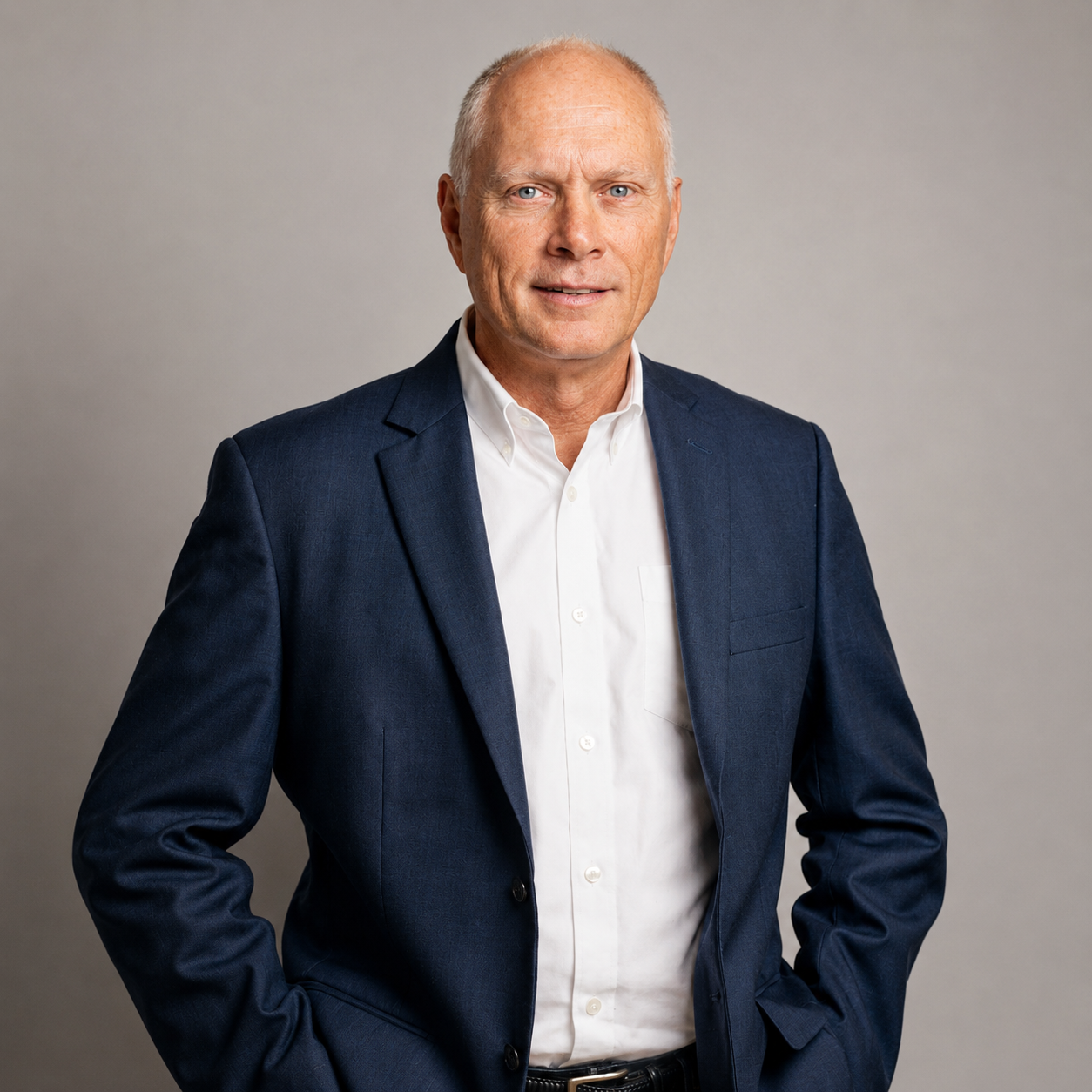 Studio portrait of a man in a navy blazer and white shirt, standing against a gray background.