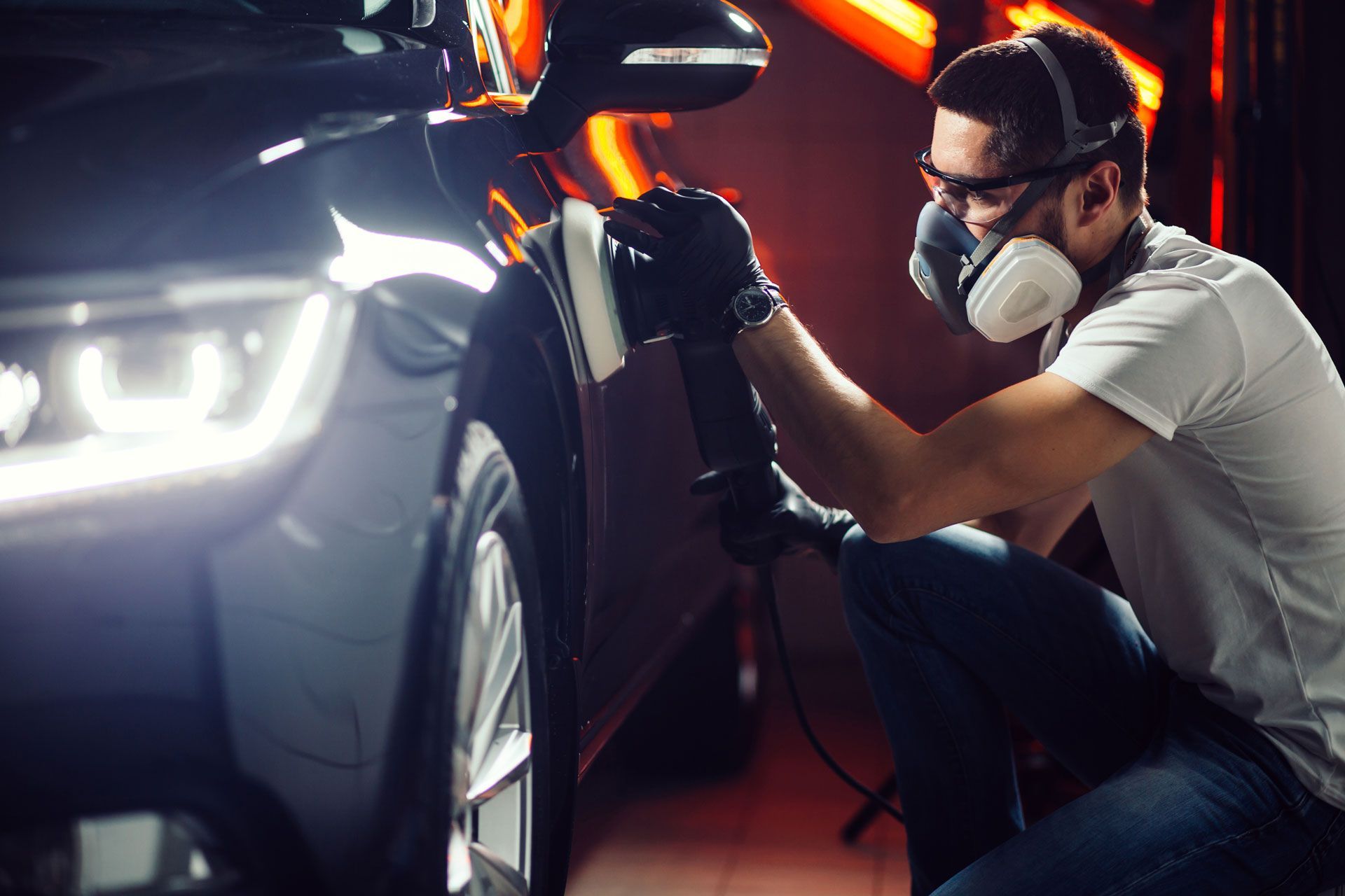 Man wearing safety gear polishing a car in a brightly lit garage.