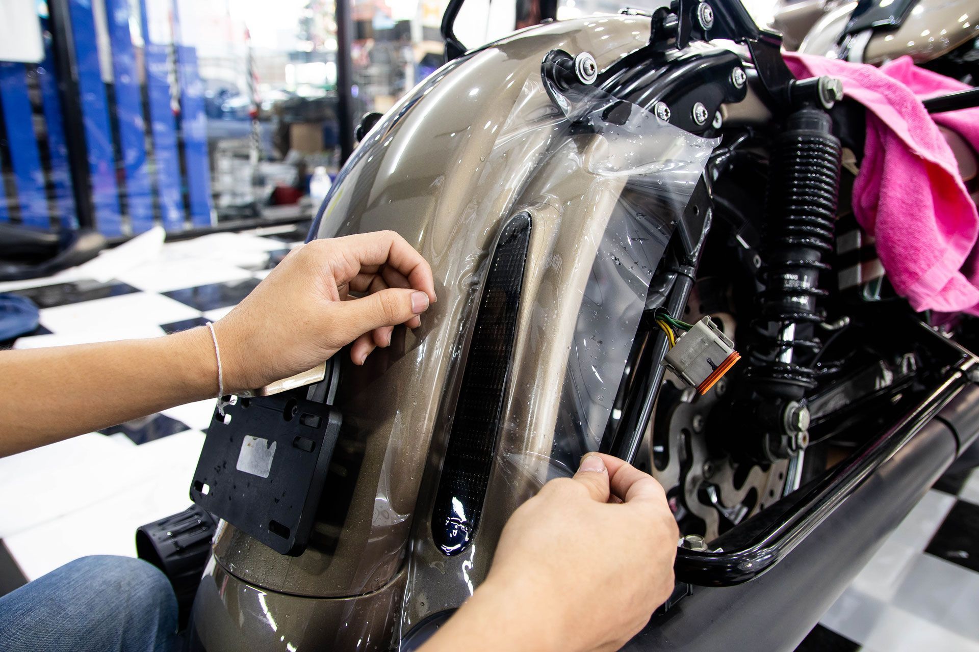 Hands installing clear film on a motorcycle fender. Setting is indoors with checkerboard floor.