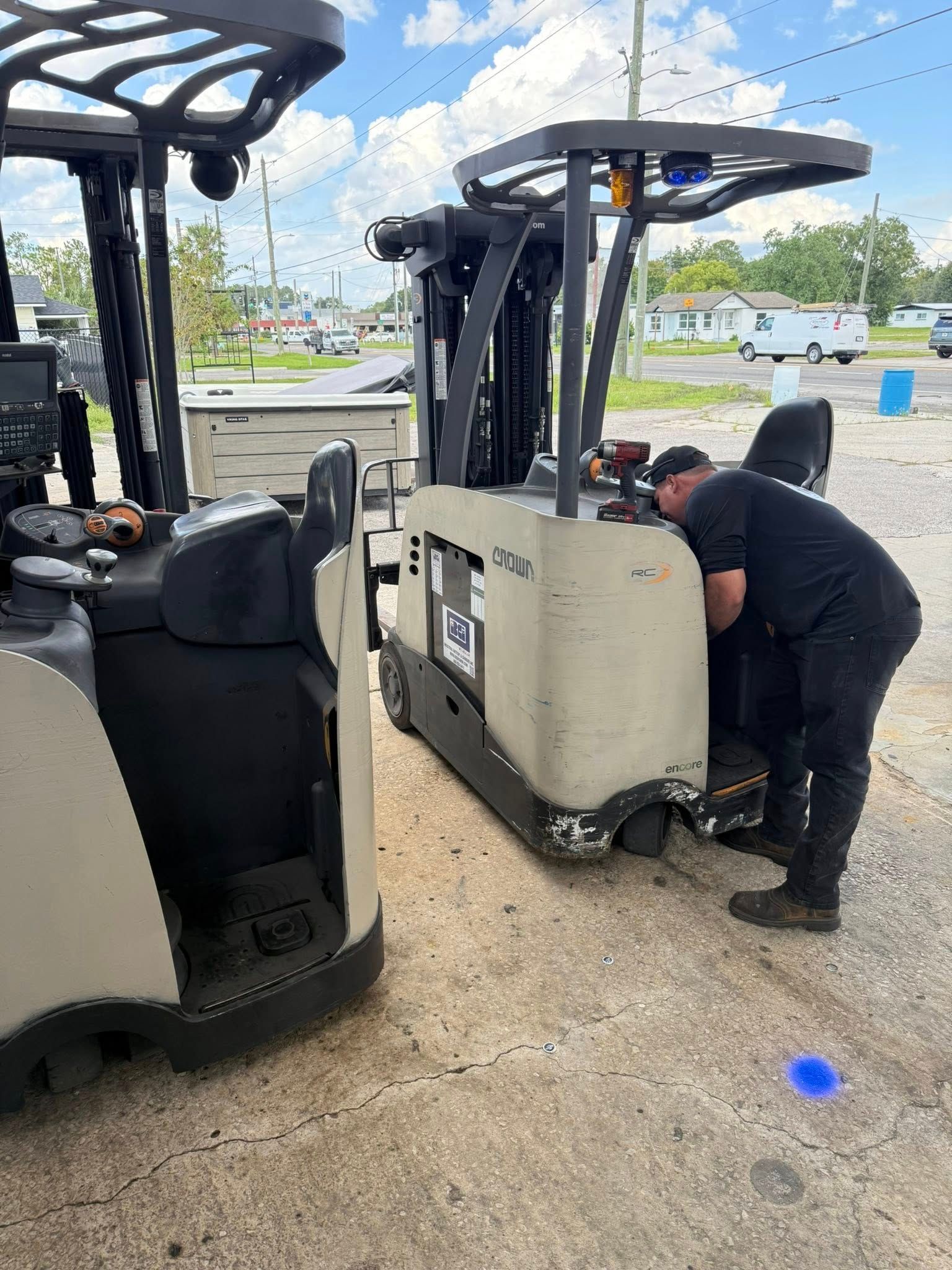 A person in a black uniform performs maintenance on a cream-colored reach forklift parked outside on a paved lot.