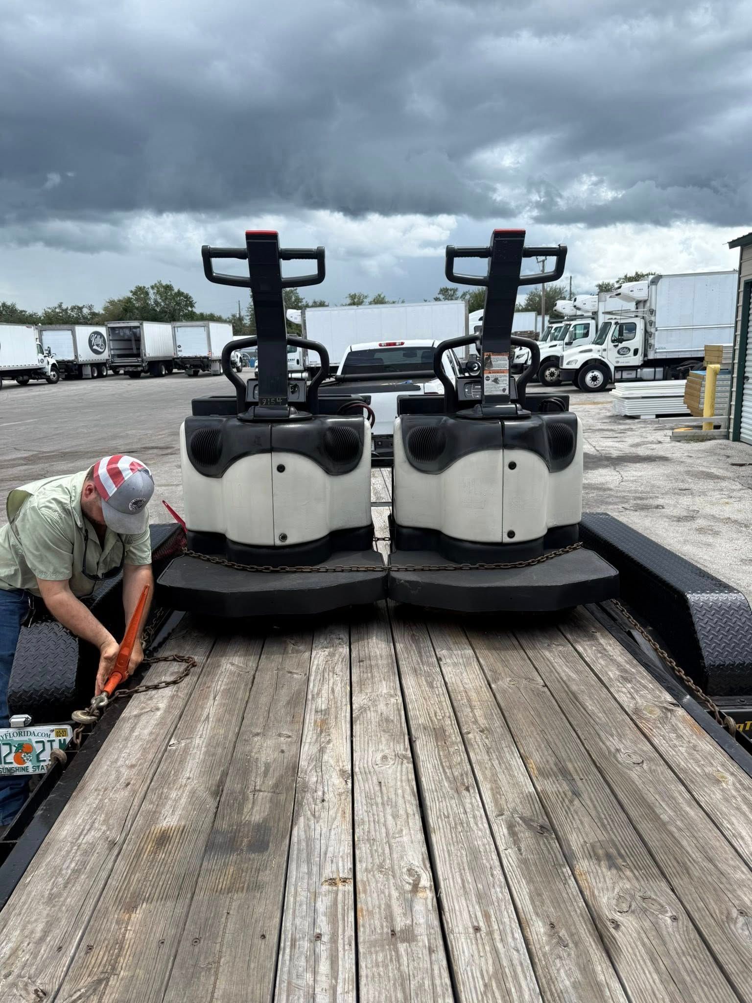 A worker secures two large, white and black industrial electric pallet jacks onto a wooden flatbed trailer.