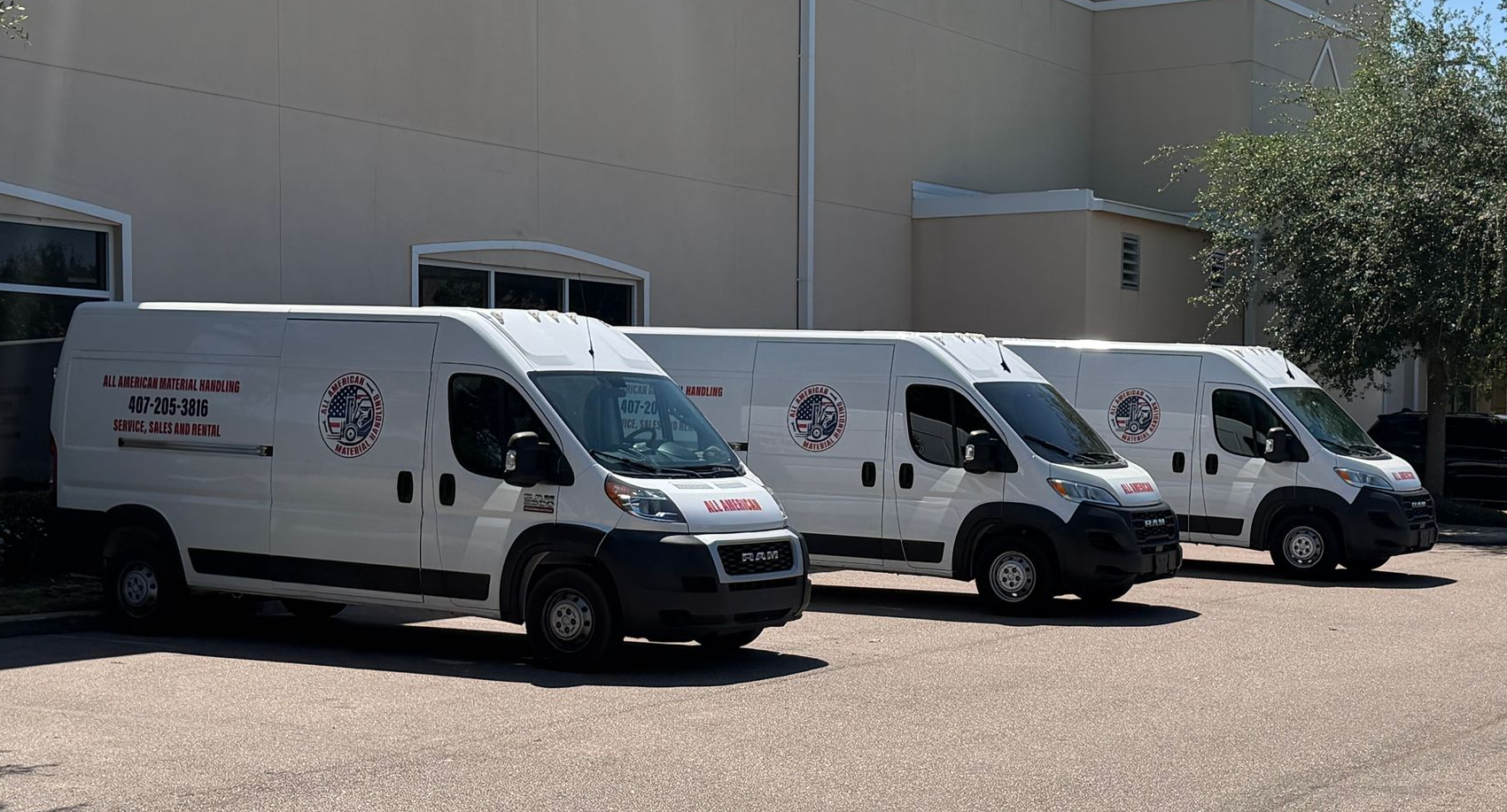 Three white delivery vans parked in a lot beside a beige industrial building.