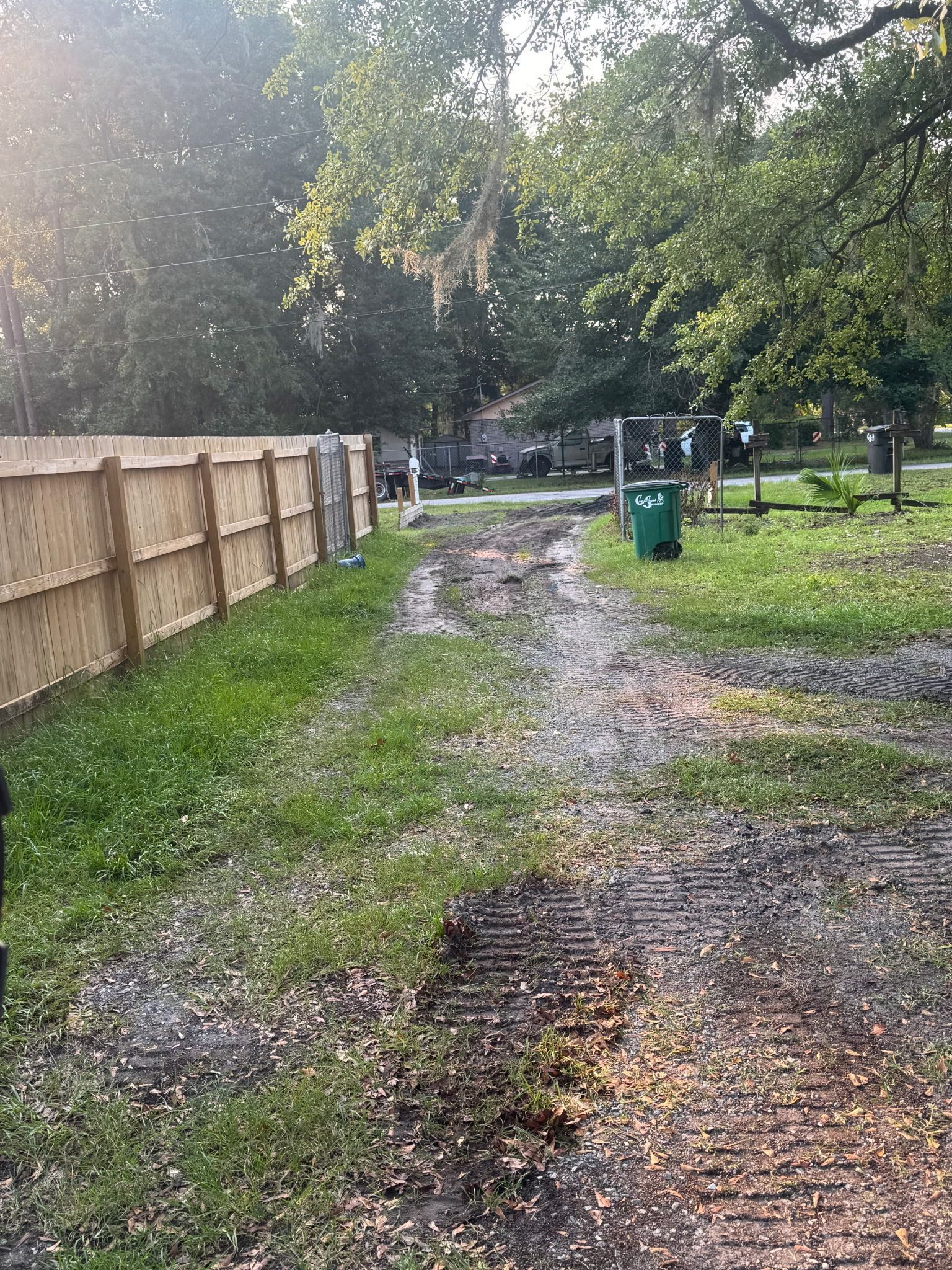 Gravel path beside a wooden fence and grassy area with trees, lit by sunlight.