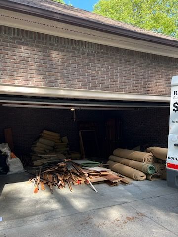 An open garage filled with piles of debris, wooden planks, and rolled-up carpet, with a utility truck parked on the right.