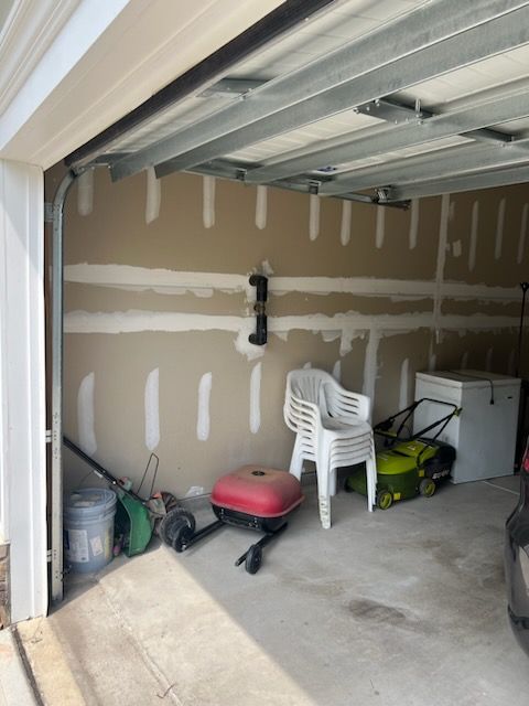 The interior of a garage with unfinished drywall walls, a white chair, a small red grill, a lawn mower, and a freezer.