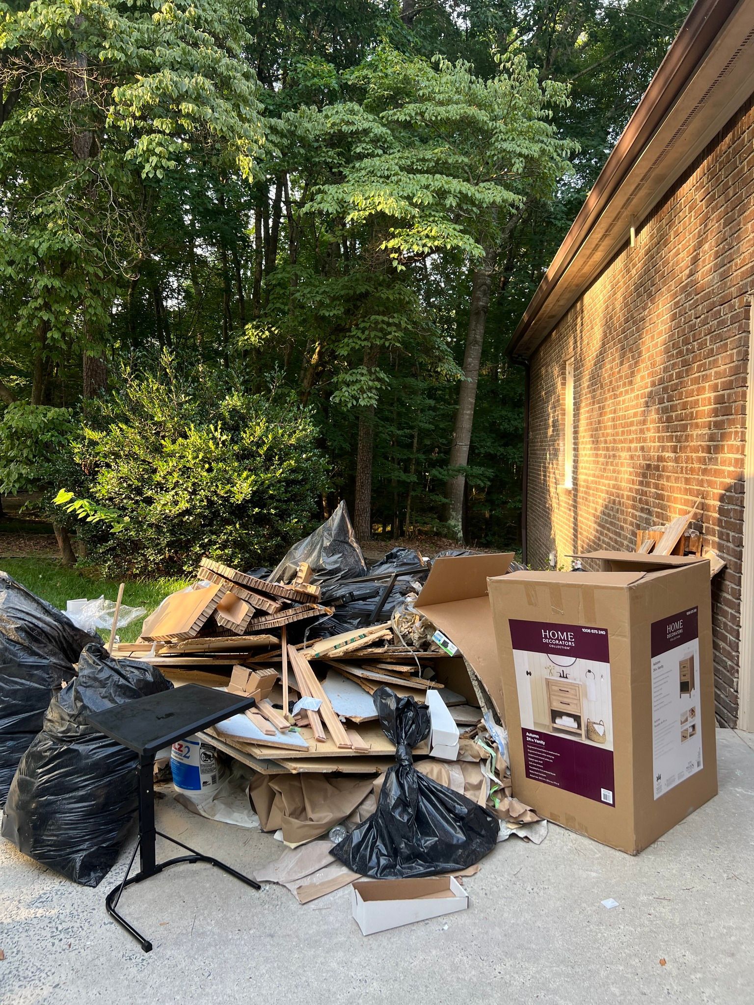 A pile of trash, cardboard boxes, and a portable desk stand sits outside next to a brick building and forest trees.