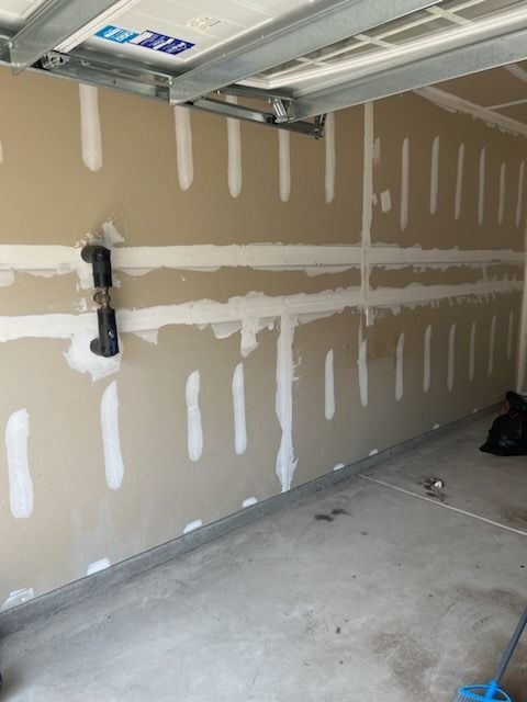 An unfinished garage interior with tan drywall featuring visible white taped joints and a black wall-mounted fixture.