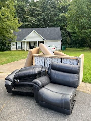 Black leather sectional and beige couch dumped in a driveway in front of a house with trees behind