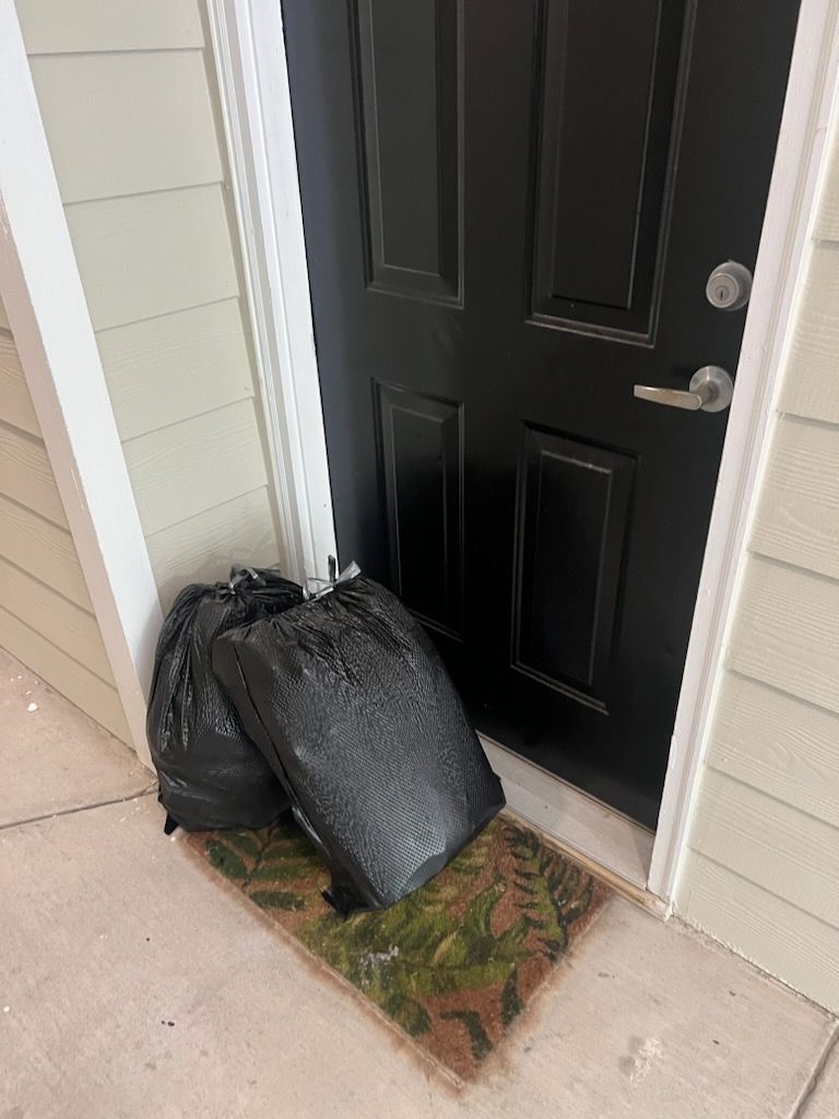 Two black trash bags sitting on a doormat in front of a dark, closed residential door.