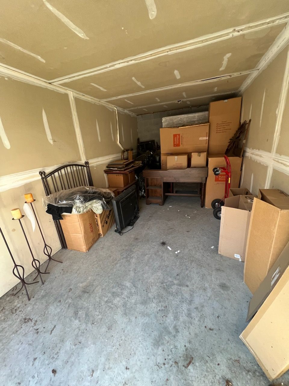 A cluttered storage unit containing several cardboard boxes, a wooden desk, a bed headboard, and tall metal candle holders.