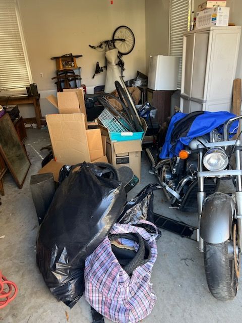 A cluttered garage with a motorcycle under a blue cover, stacked cardboard boxes, bags, and a bicycle mounted on the wall.