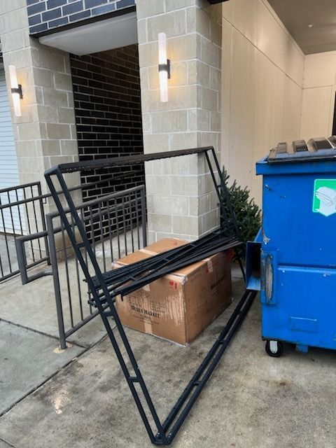 A black metal frame leaning against a brick building next to a cardboard box and a blue dumpster.