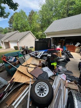 A driveway filled with clutter, including a spare tire, pieces of wood, a sofa, and other debris near an open garage.