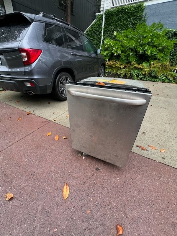 A stainless steel dishwasher stands on a sidewalk next to a grey SUV, waiting to be picked up for trash or recycling.