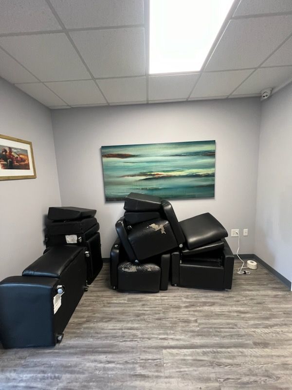 A room containing a stack of black leather chairs and a horizontal landscape painting on the grey wall.