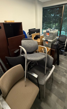 A cluttered office space featuring a stack of several gray and brown office chairs, storage bins, and furniture.