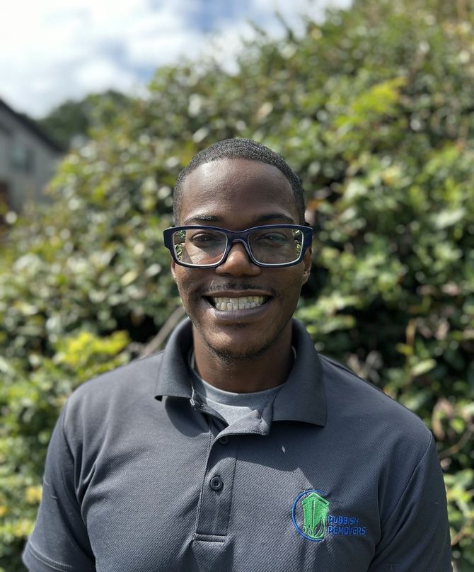 A person wearing glasses and a gray polo shirt smiling in front of a leafy green bush.