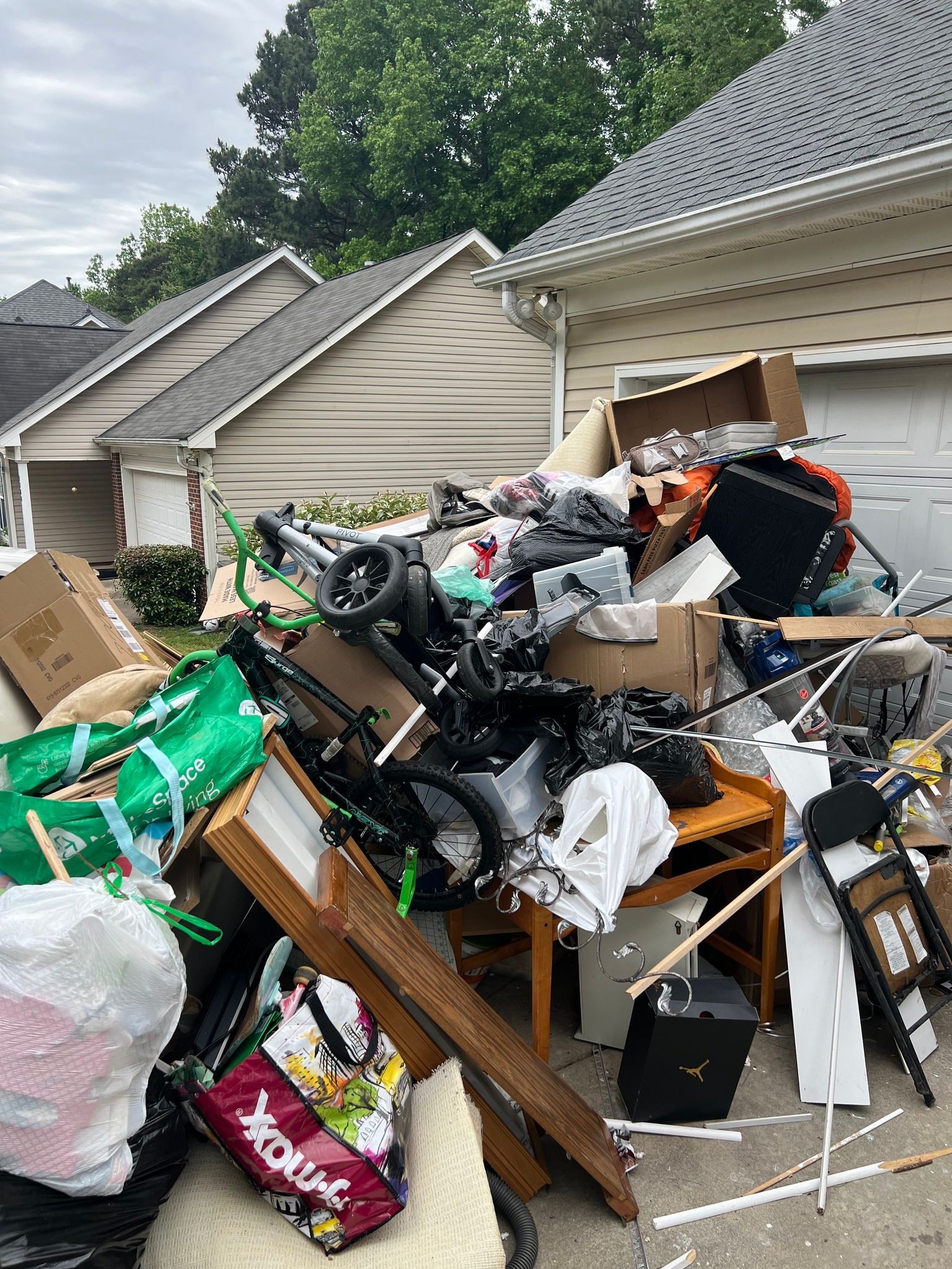 A large pile of household junk, boxes, and furniture sits in a driveway in front of a suburban garage.