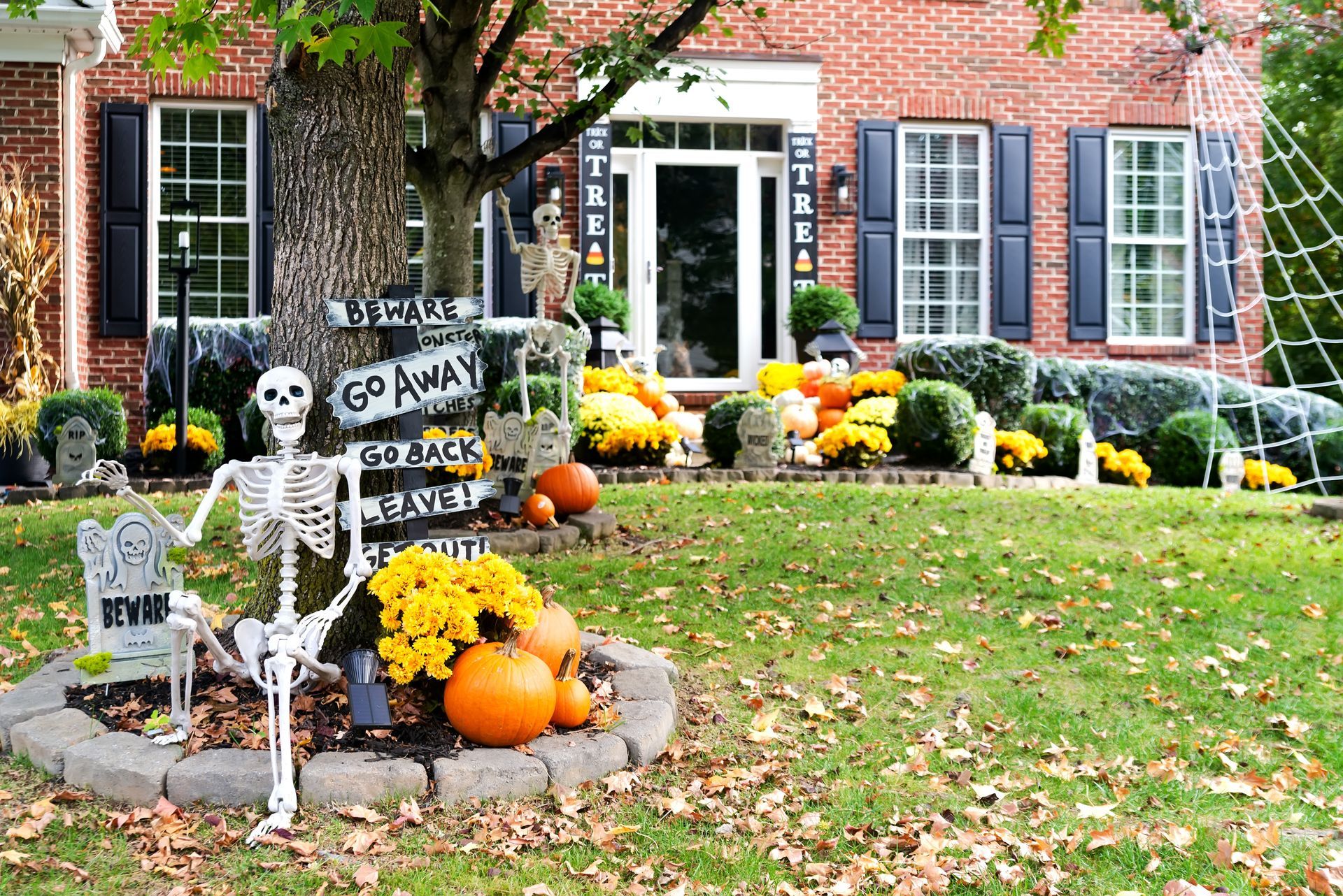 Halloween-decorated brick house with skeletons, pumpkins, and autumn flowers on the lawn