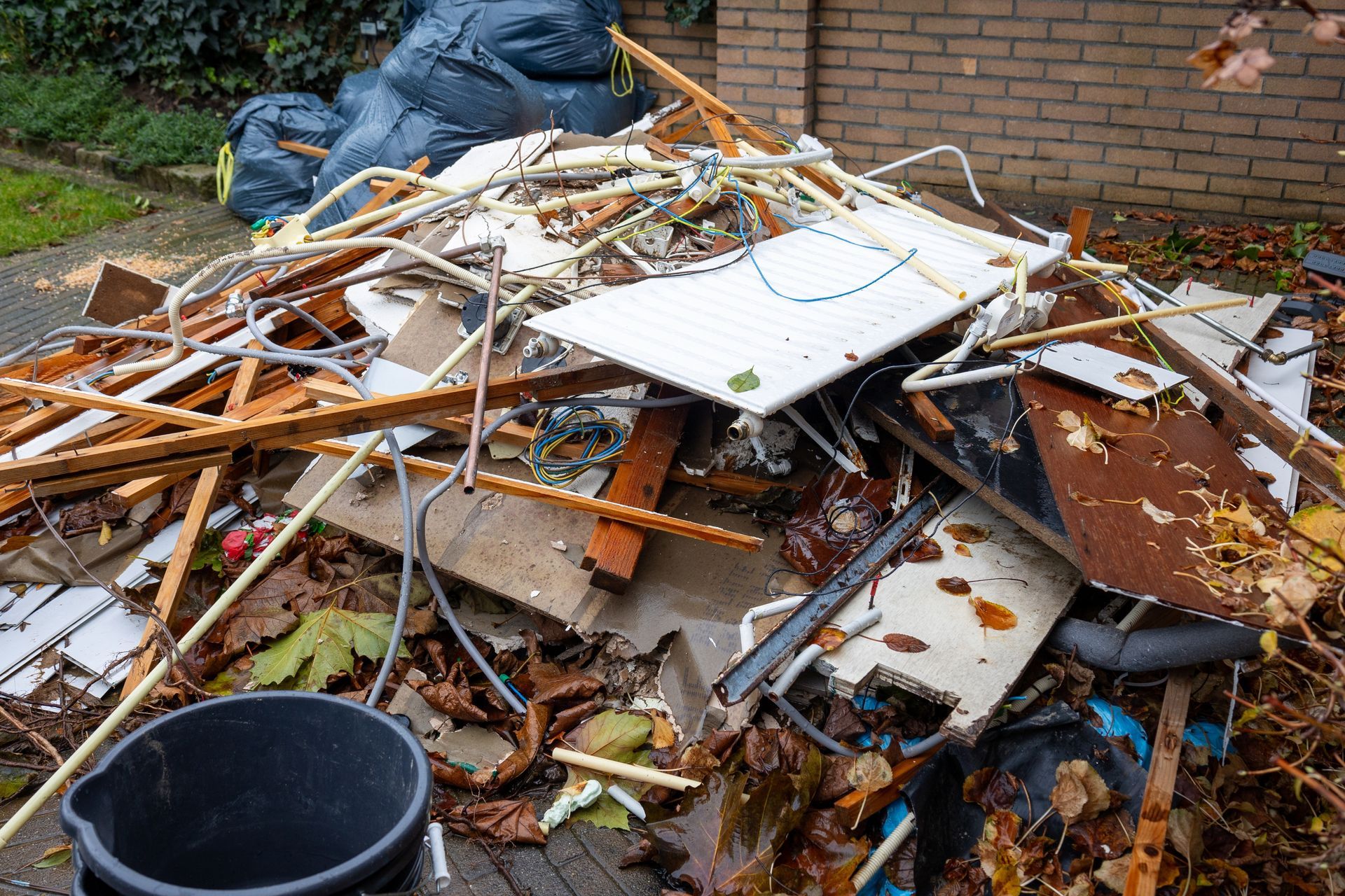 A large, messy pile of construction debris, scrap wood, metal pipes, and trash bags sitting next to a brick wall.