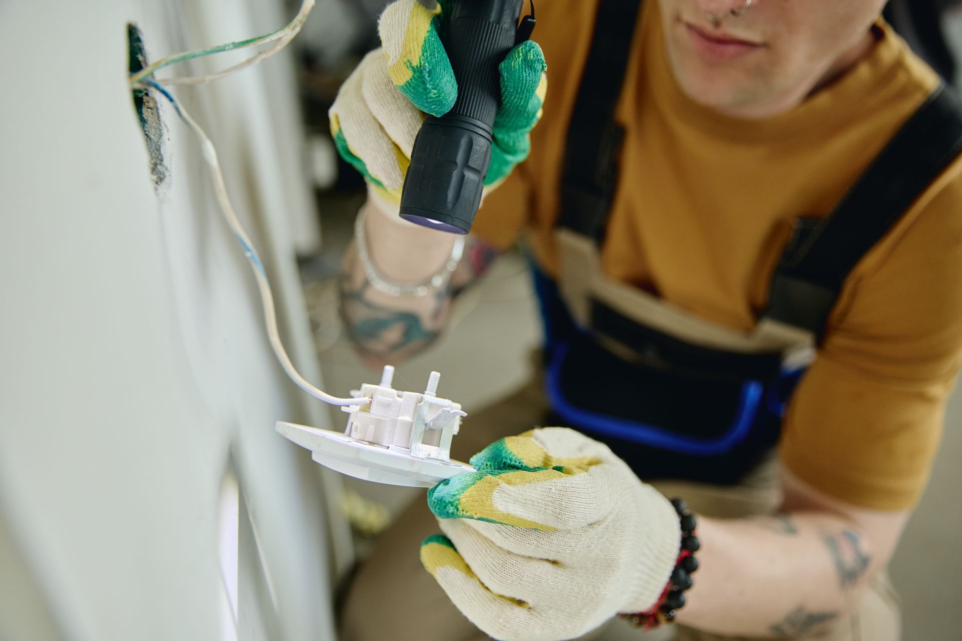 Electrician in work gloves uses a flashlight while installing an outlet in a wall.