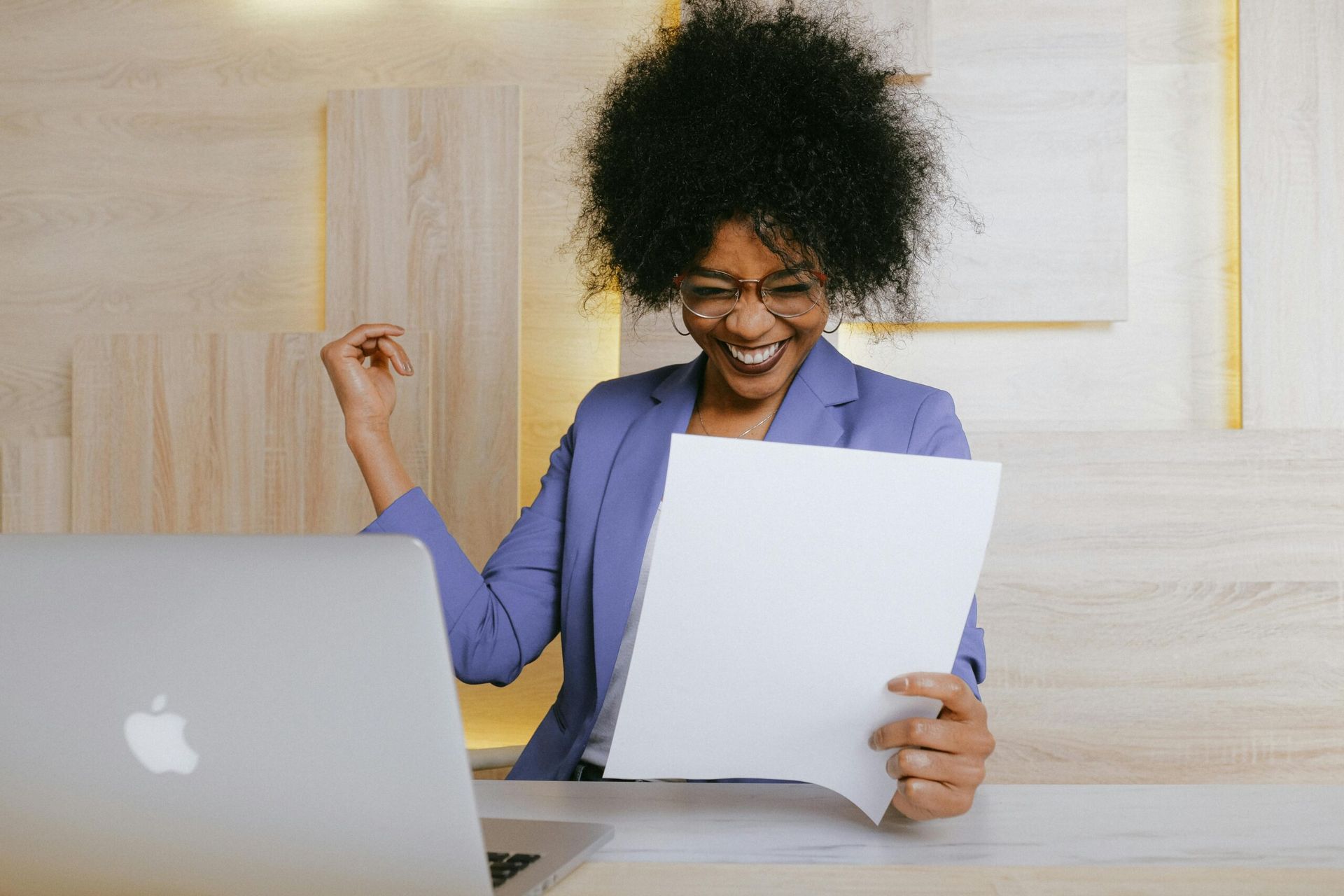 Woman in blue blazer smiles, raises arm, and looks at document near laptop.