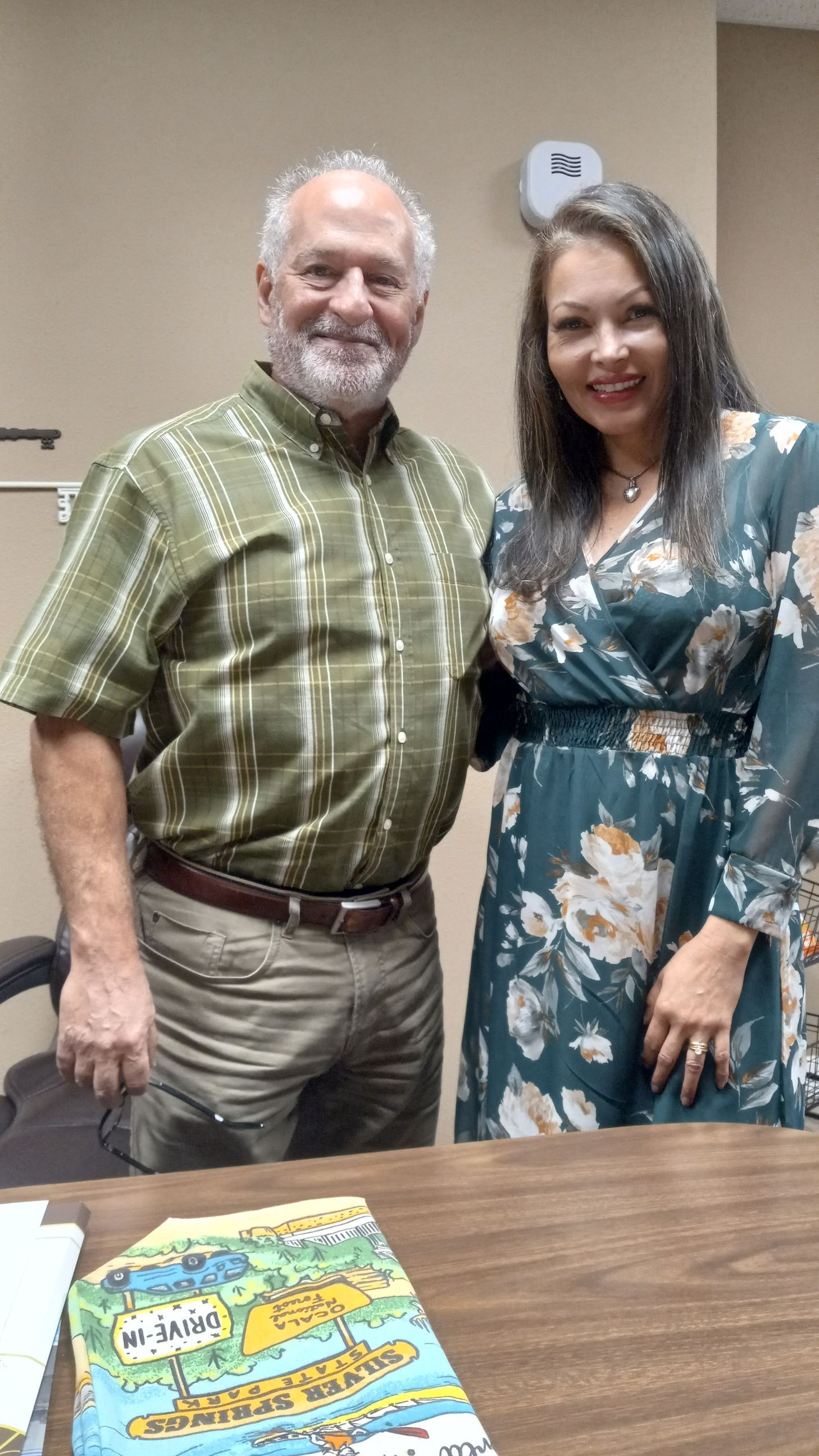 Man and woman smiling together, indoors. Woman in floral dress, man in patterned shirt.