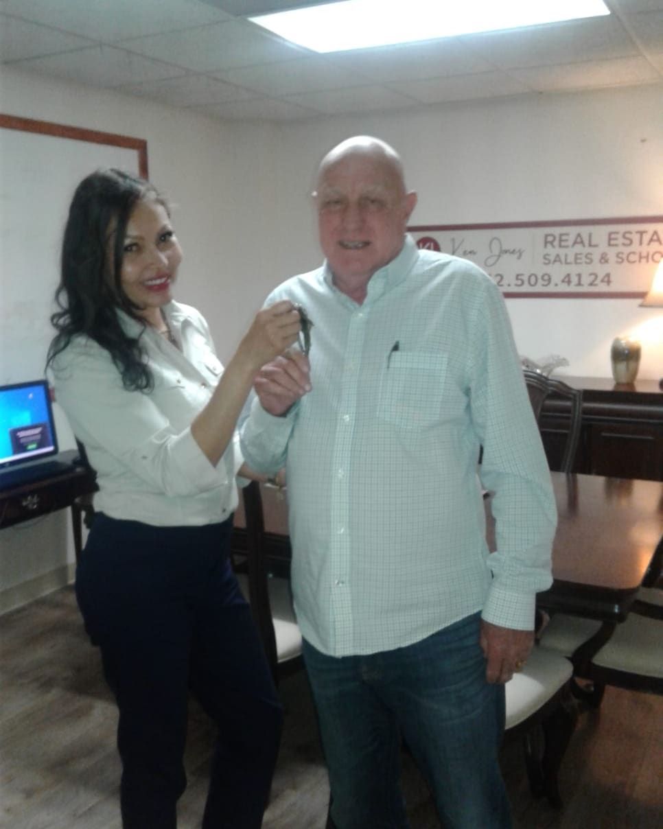 Woman handing keys to a man in an office. They are smiling. A Real Estate sign is in the background.
