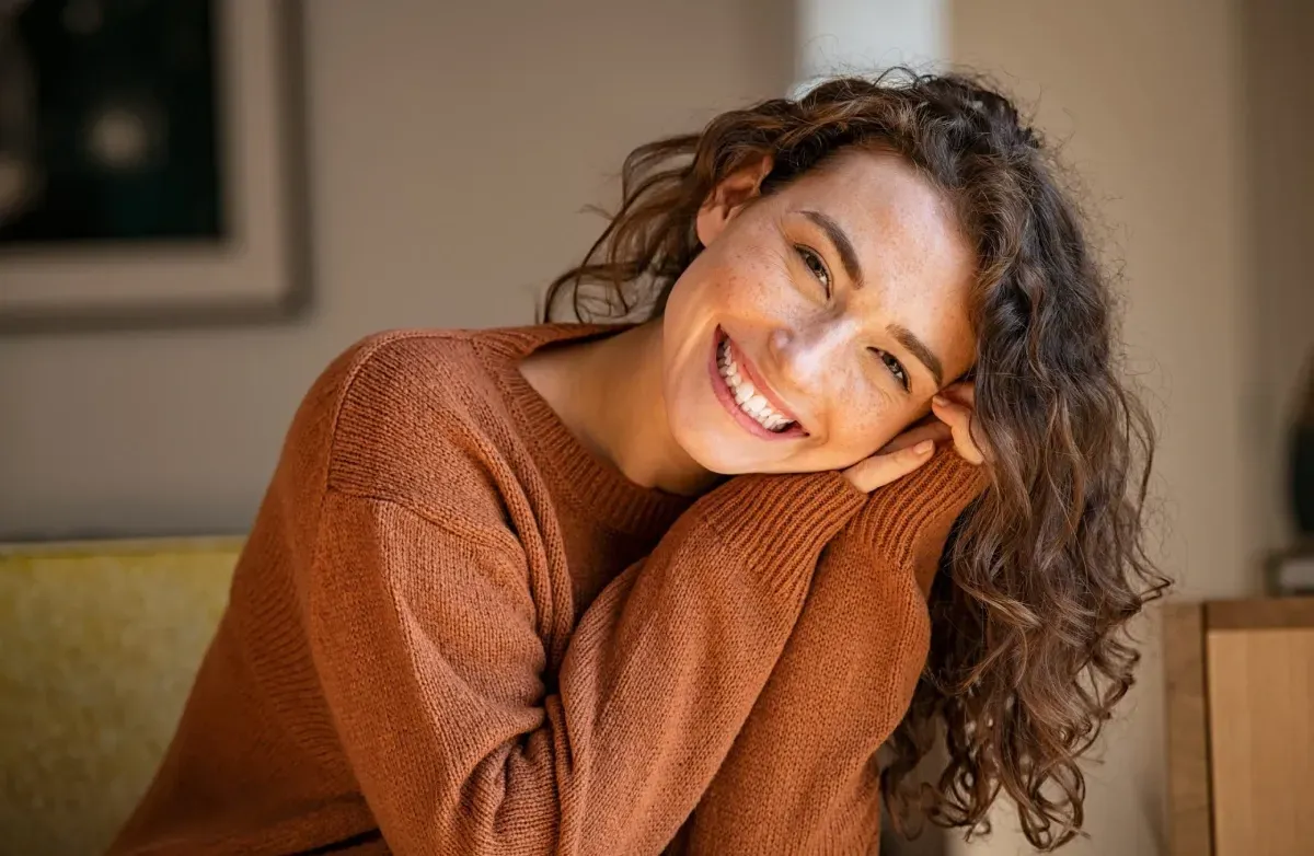 Smiling person in a cozy brown sweater, leaning their head on folded hands indoors.