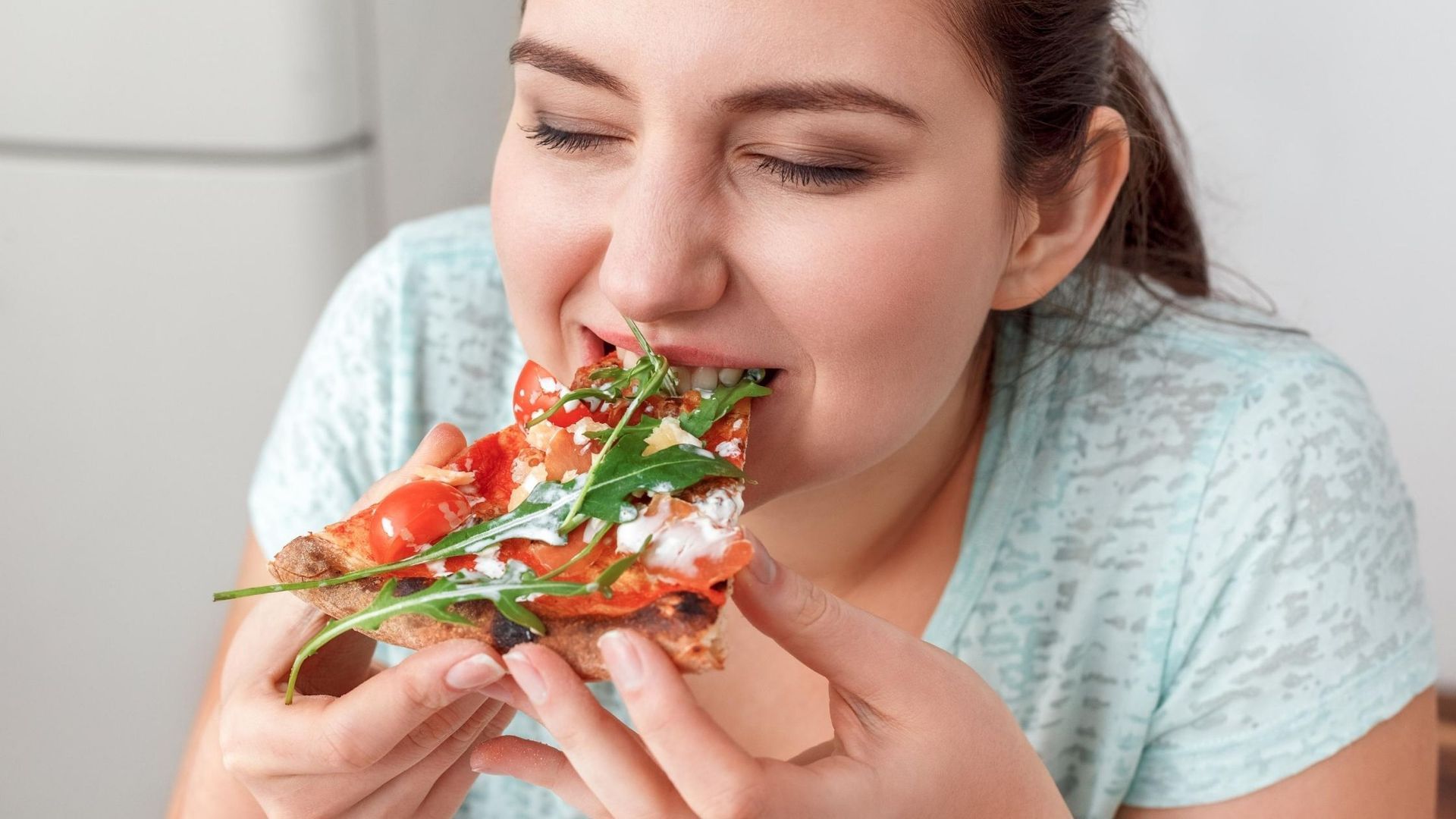 A person with their eyes closed enjoys a slice of pizza topped with fresh arugula and tomatoes.
