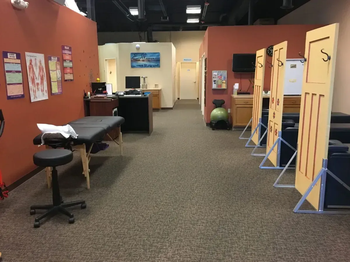 Interior of a clinic waiting area with exam tables, stools, and privacy partitions along a carpeted hallway.