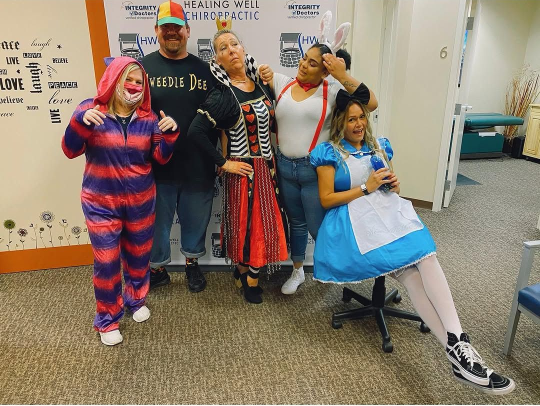 Group in colorful costumes posing indoors, one seated in a blue dress holding a prop sign.