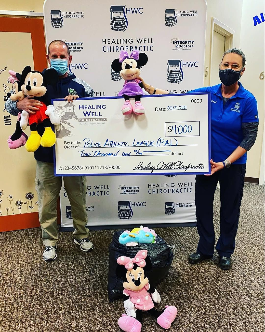 Two masked people hold a giant check beside Mickey and Minnie Mouse at a charity event banner.