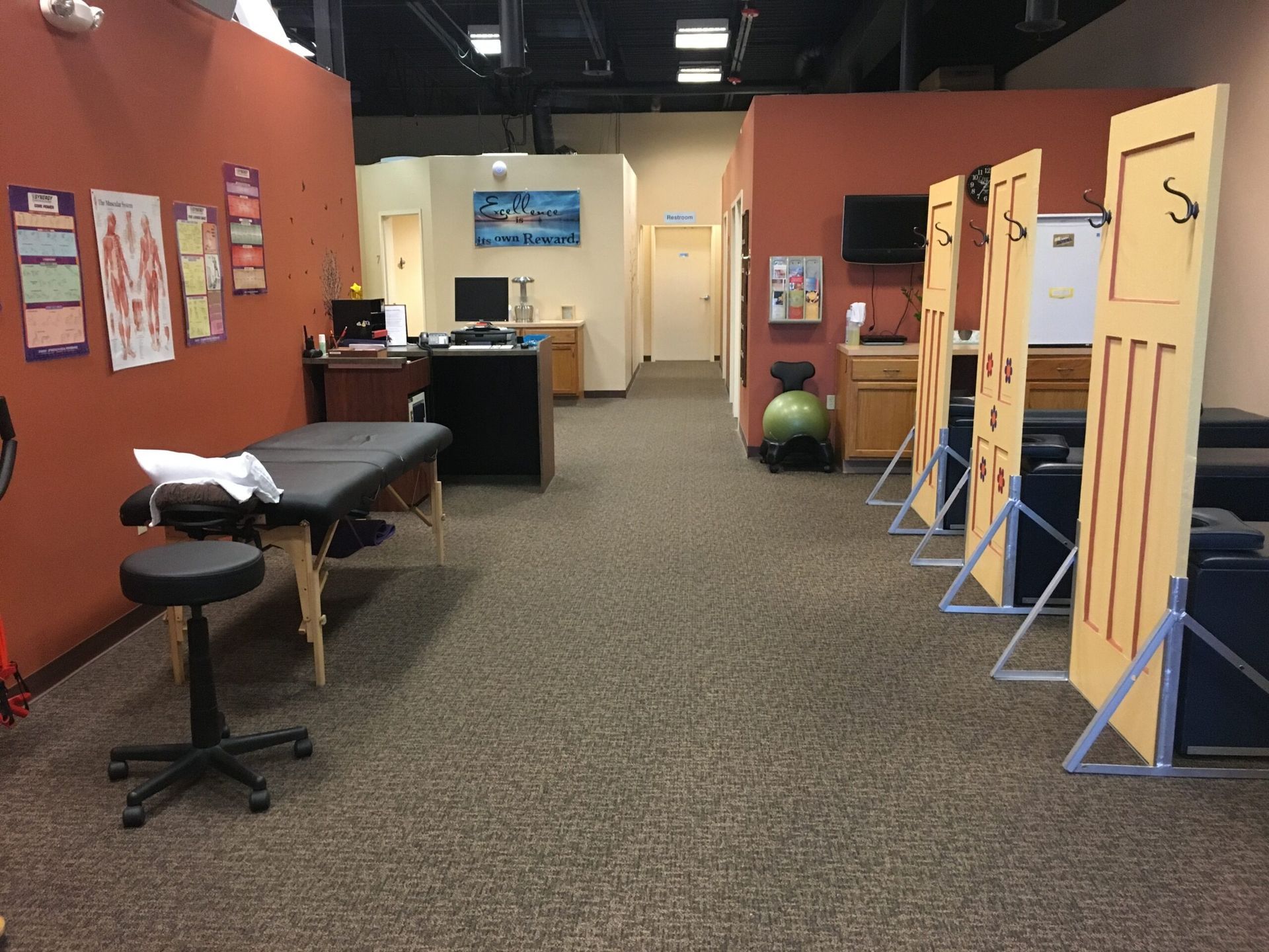 Interior of a clinic waiting area with exam tables, stools, and privacy partitions along a carpeted hallway.