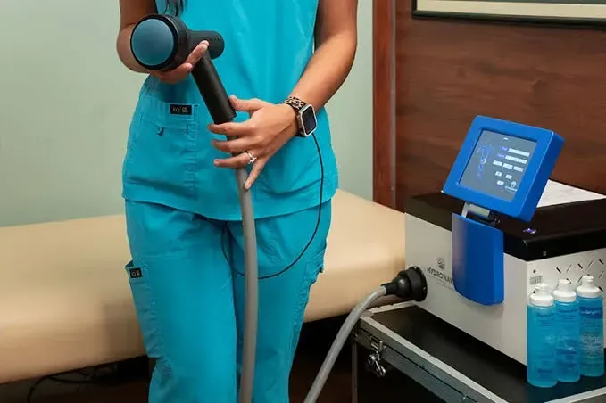 Person in teal scrubs holding a handheld medical device beside a clinic machine and water bottles