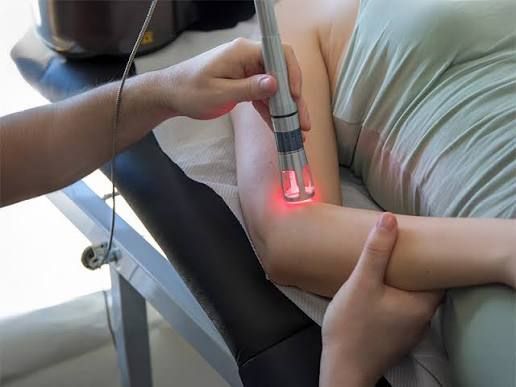 A person receiving laser treatment on an arm while seated in a clinic chair.