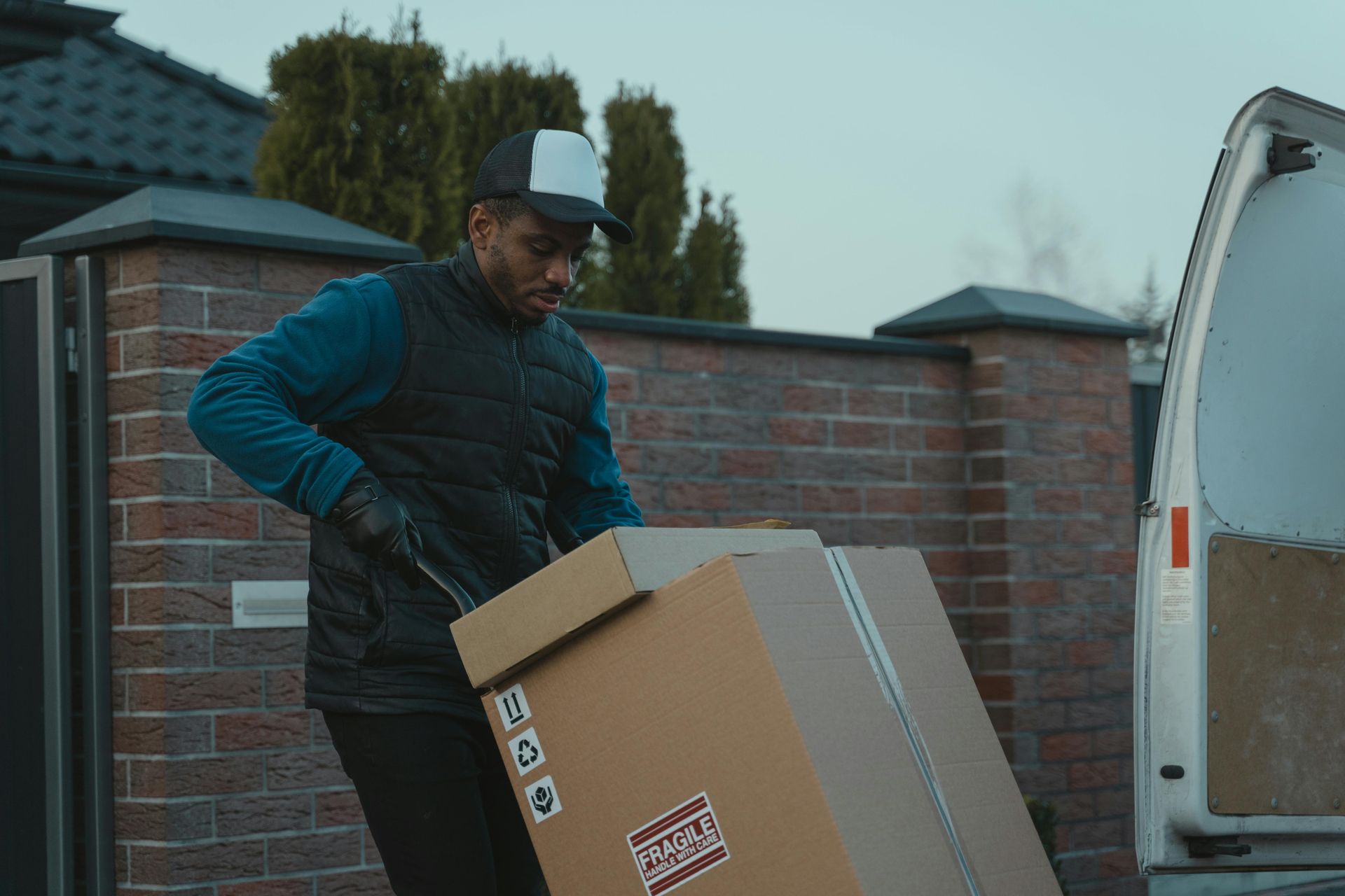 Zed Logistics delivery driver lifting a large cardboard box beside a white van and brick wall