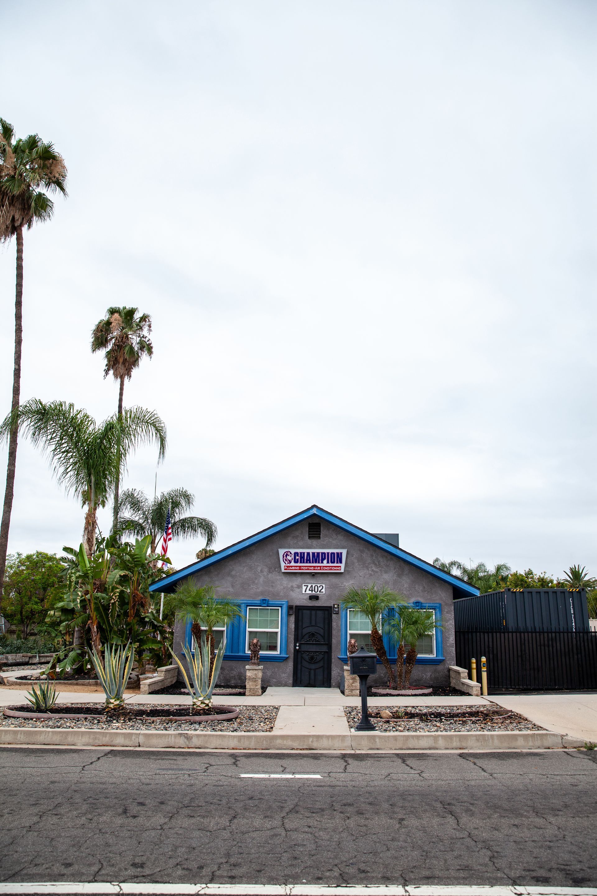 Small blue storefront with a sign, palm trees, and a sidewalk beside a road under an overcast sky