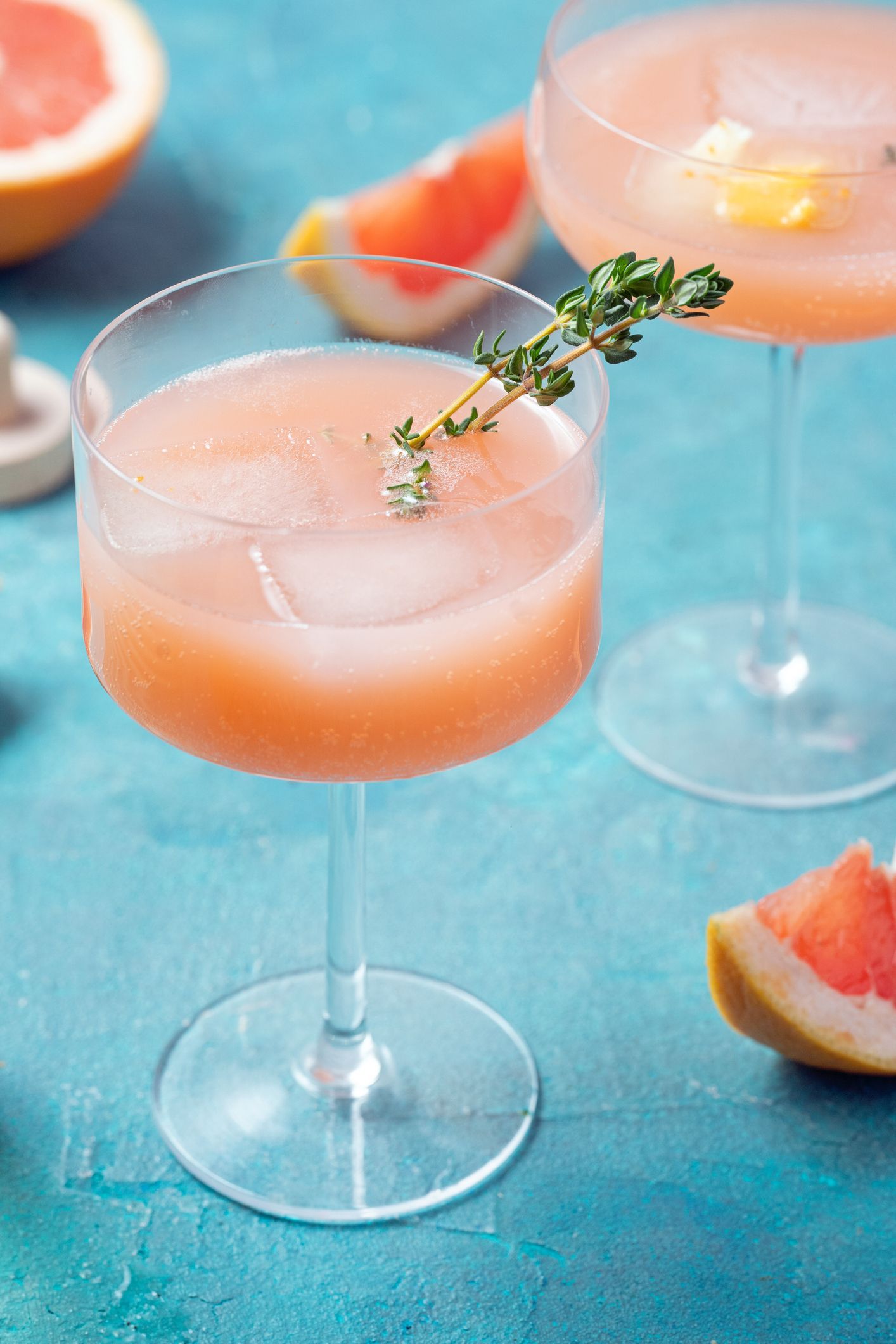 Two pink grapefruit cocktails garnished with thyme, on a blue surface.