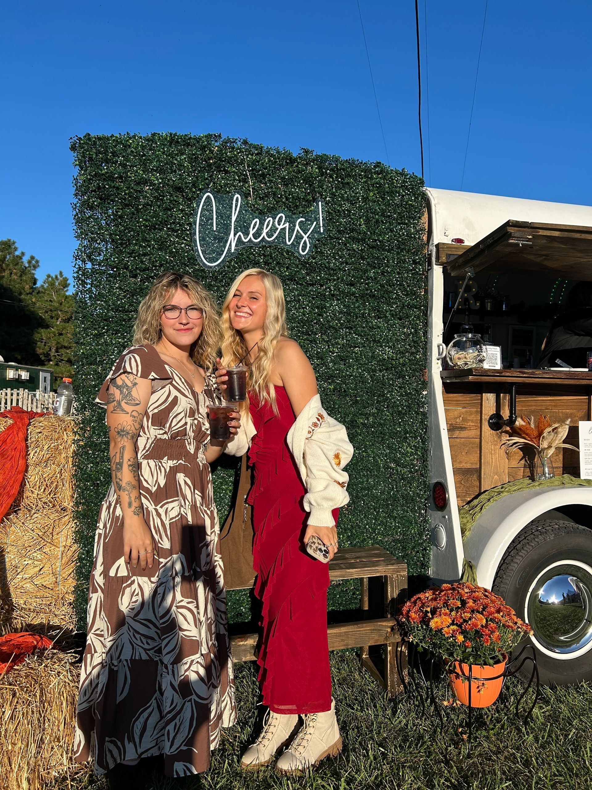 Two women at an outdoor event, one in red and one in patterned dress, pose near a 