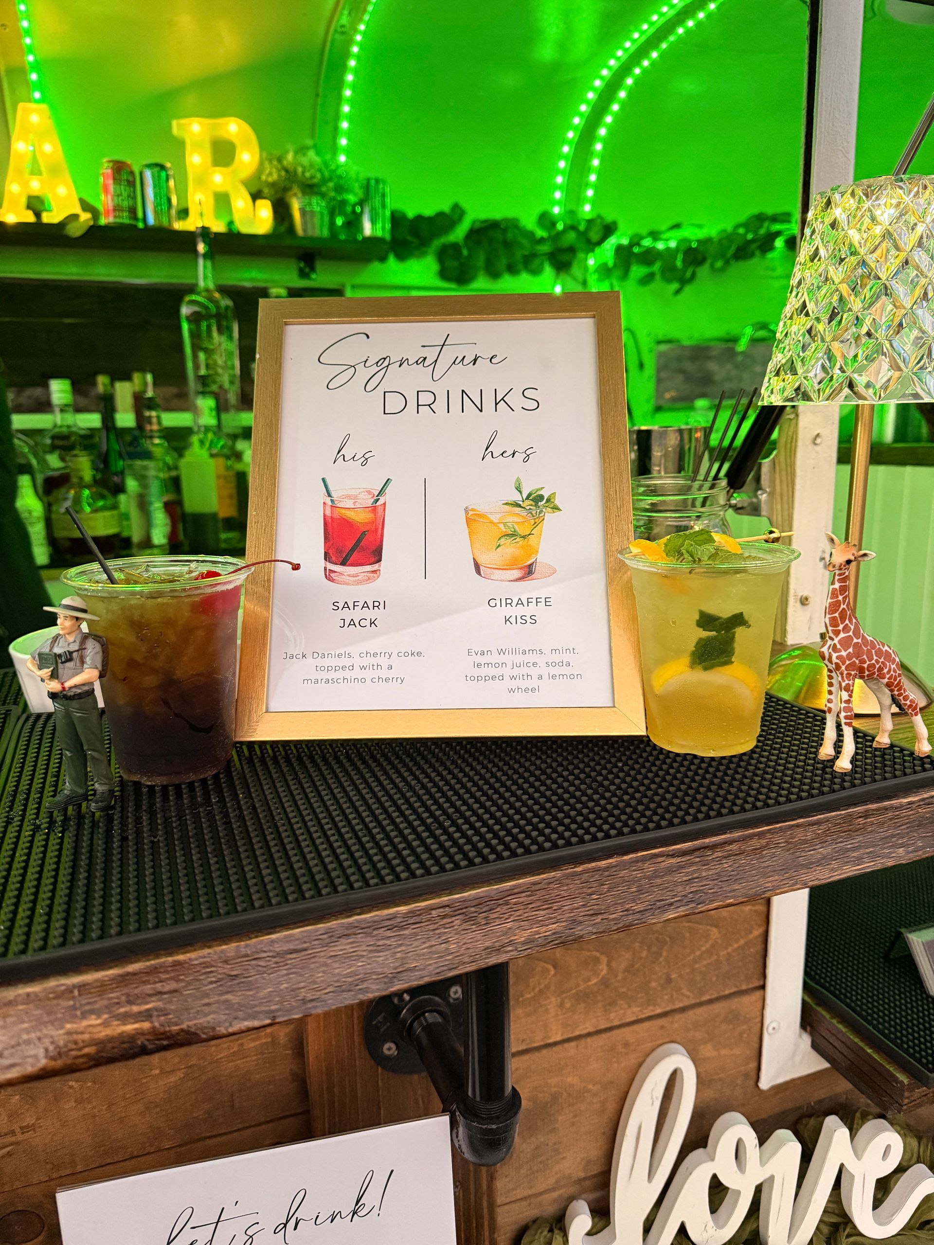 Bar setup with signature drink sign and two cocktails; green neon lights; wooden bar with 