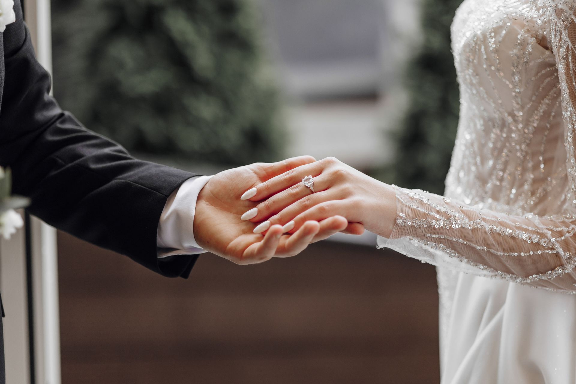 Bride and groom holding hands, wedding ring visible, white dress, black suit, outdoor setting.