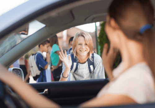 Smiling woman waving from a car window at a school pickup line