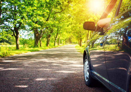 Car parked on a sunlit tree-lined road with bright green foliage and lens flare