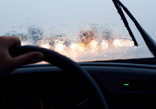 Driver’s view through a rainy windshield at sunset, with blurred headlights ahead and a hand on the steering wheel