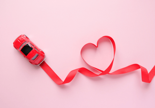 Red toy car pulling a red ribbon shaped like a heart on a pink background