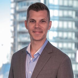 Professional headshot of a smiling man in a blazer against a blurred office building background