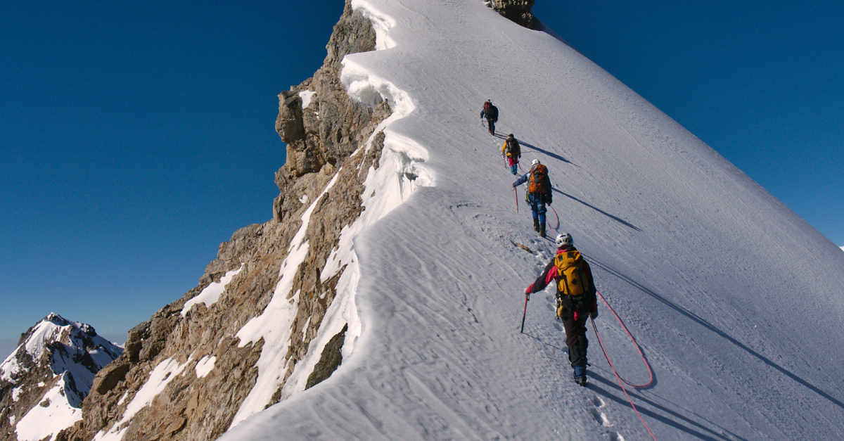Mountaineers climbing a snowy ridge with ropes under a clear blue sky
